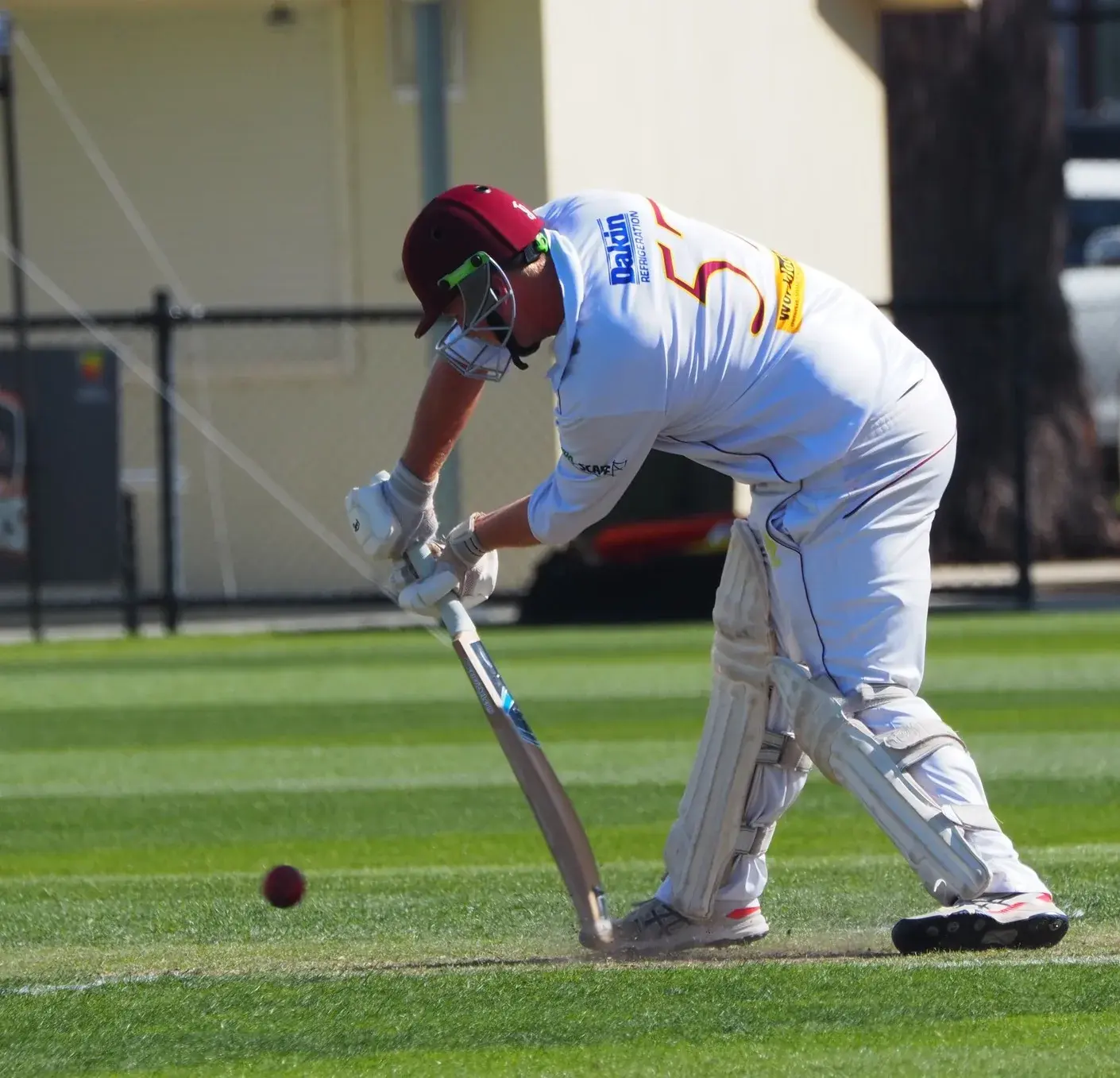 Cricketer batting on sunny field, bowler behind stumps.